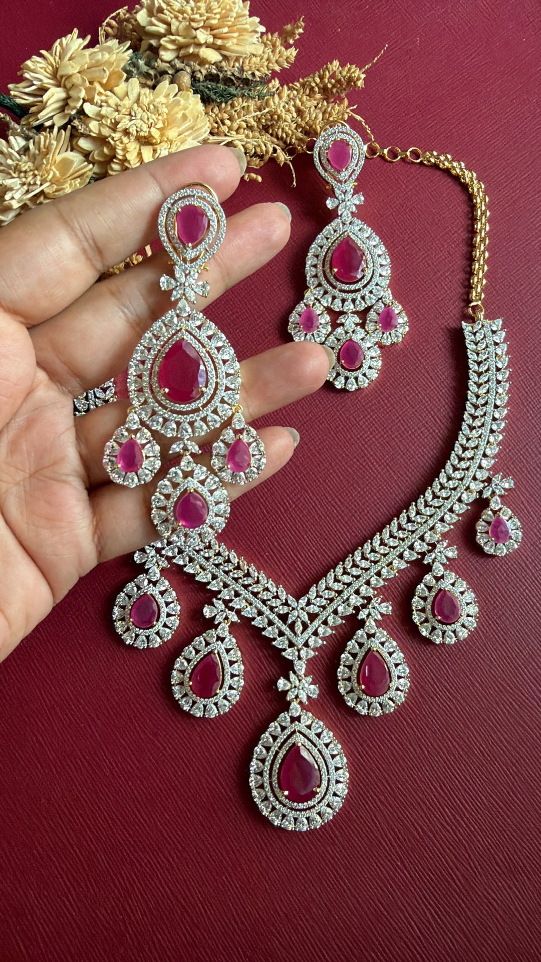 Supriya 3-inch ruby-pink American Diamond chandelier earring held in hand with matching necklace
