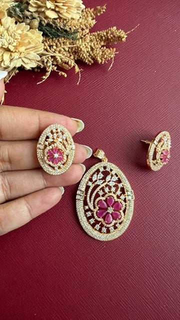 Mishka oval pendant with ruby-pink stone flower and white American Diamond spray on a gold-toned setting, with studs held in hand