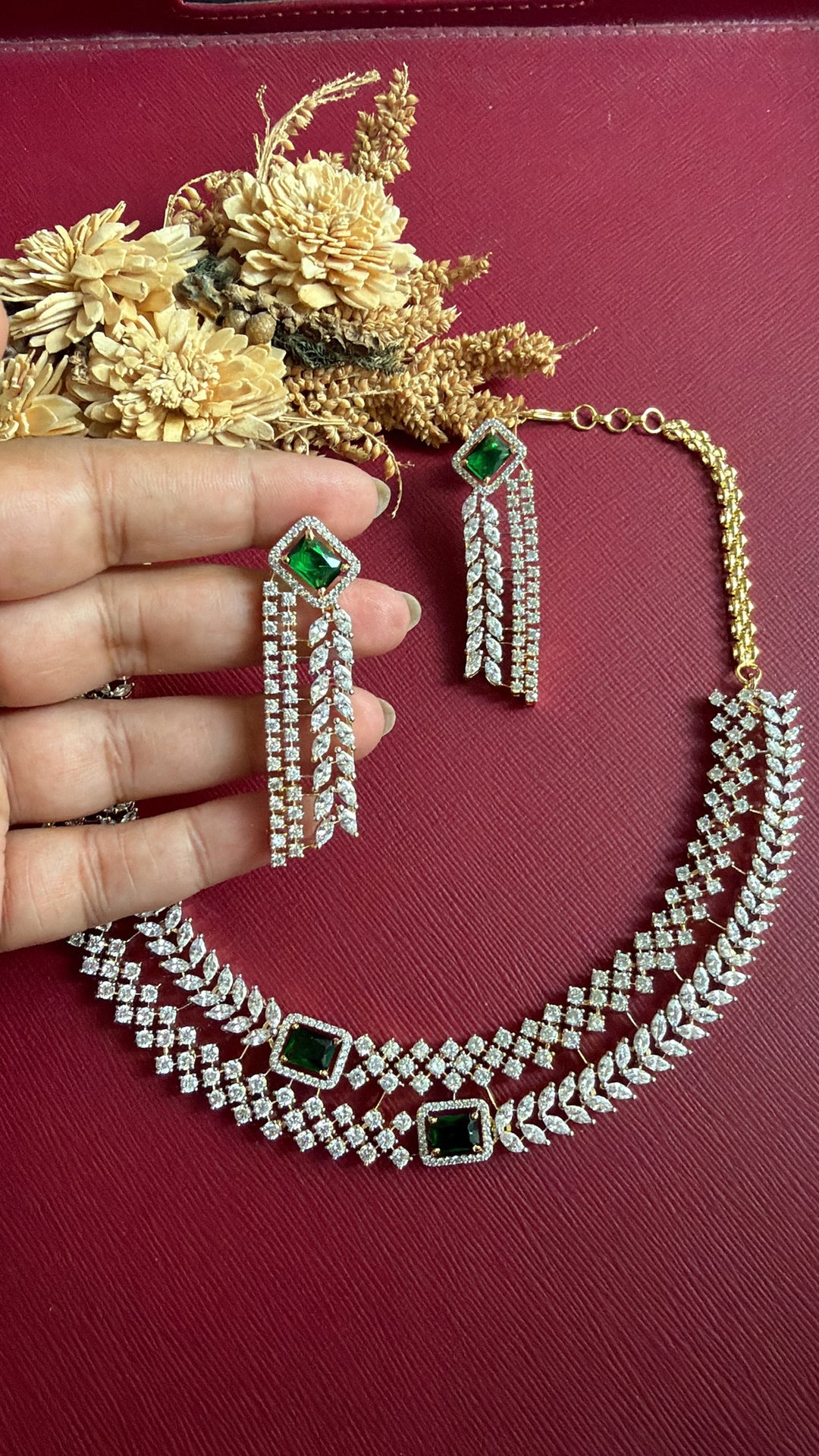 Vedika emerald-green and white American Diamond fringe earring held in hand, with the layered necklace below