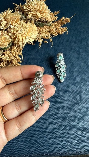 A hand holds a silver-toned leaf-shaped pendant with clear rhinestones, with a second matching pendant resting on a dark blue textured surface next to dried flowers.